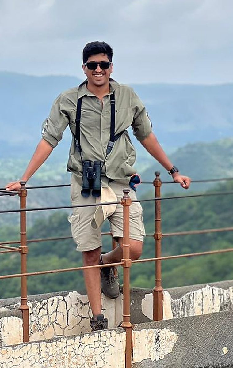 Akarsh Suresh standing on a fort rampart in a field shirt with binoculars on a chest harness, forested hills and monsoon cloud behind him