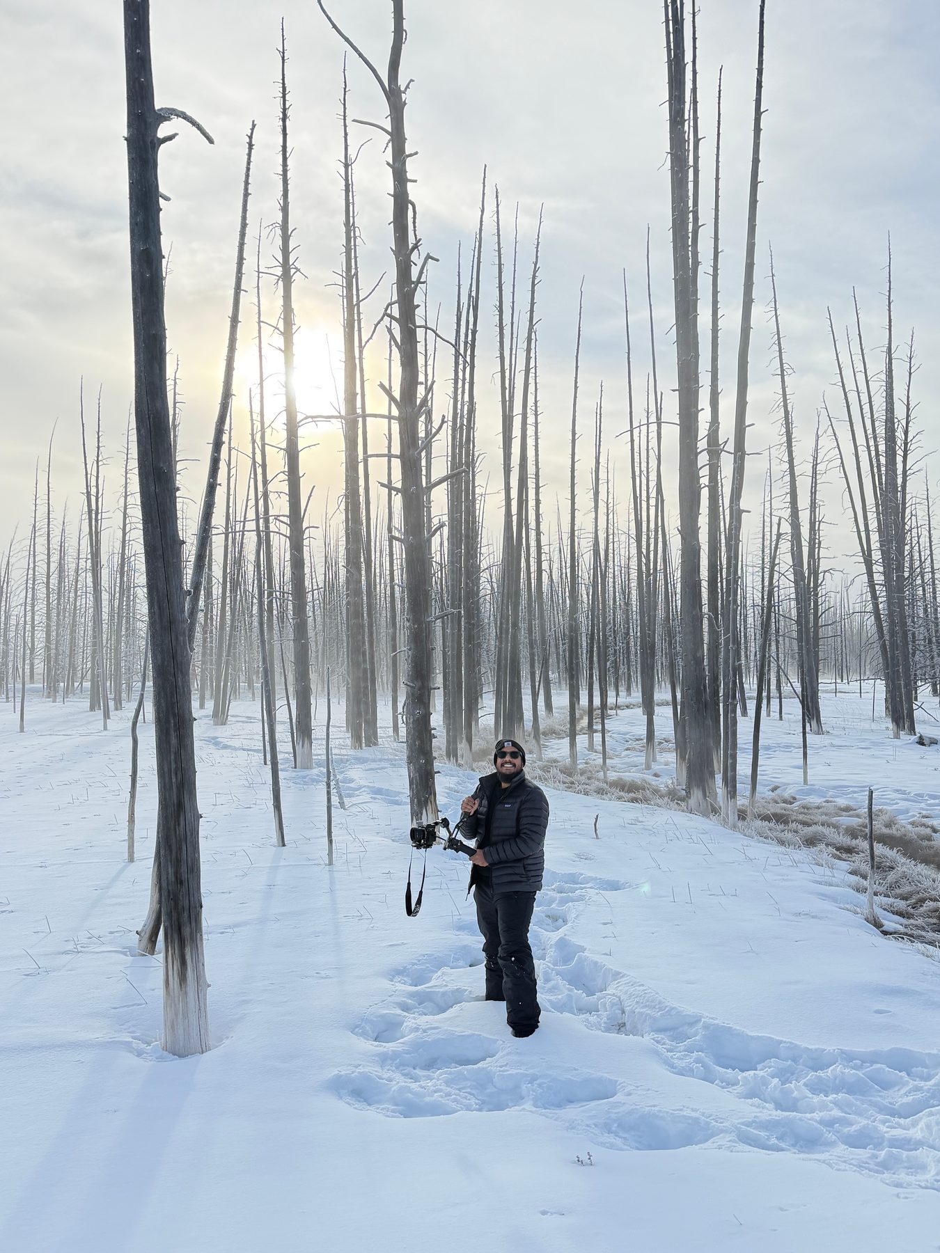 Lala lying flat in the snow behind a camera on a low tripod, photographing across a frozen lake, bare storm-killed trees and low sun beyond him