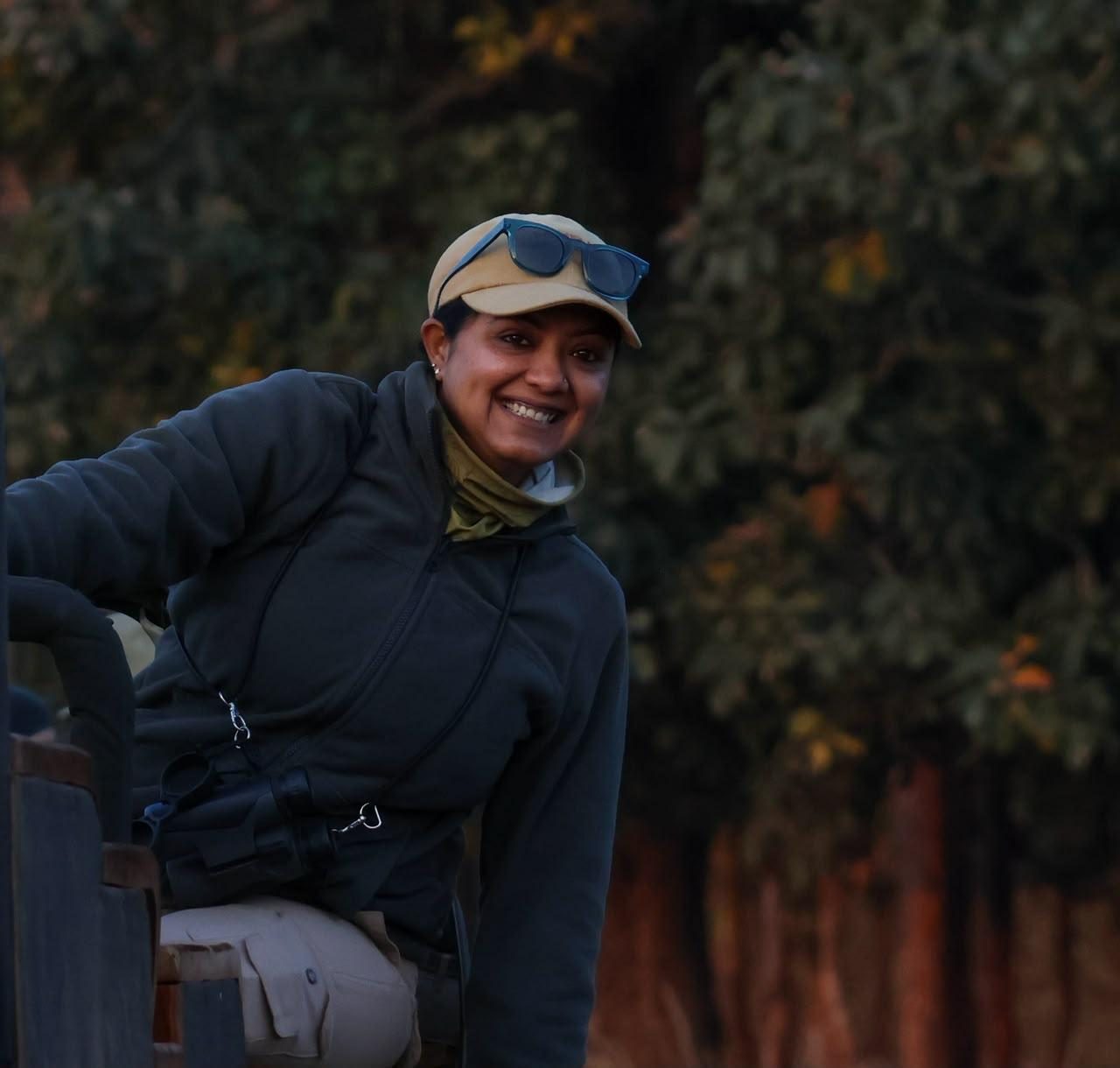 Priyanka S Murthy leaning from the back of an open safari vehicle, smiling towards the camera, binoculars on a harness and a cap pushed back, forest behind her