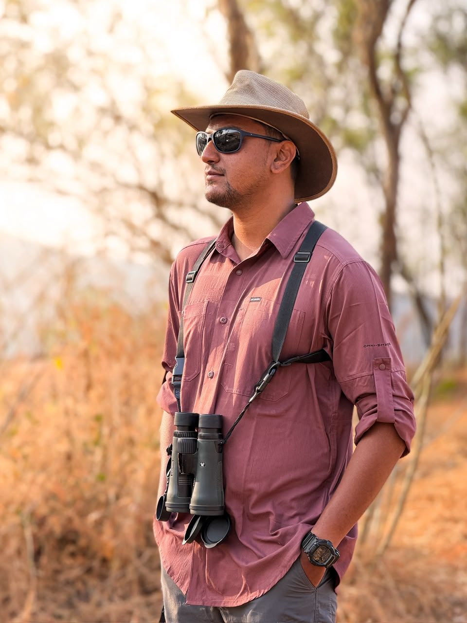 Swanand Deshpande standing in dry deciduous forest in a wide-brimmed hat and sunglasses, binoculars on a chest harness, looking off to one side